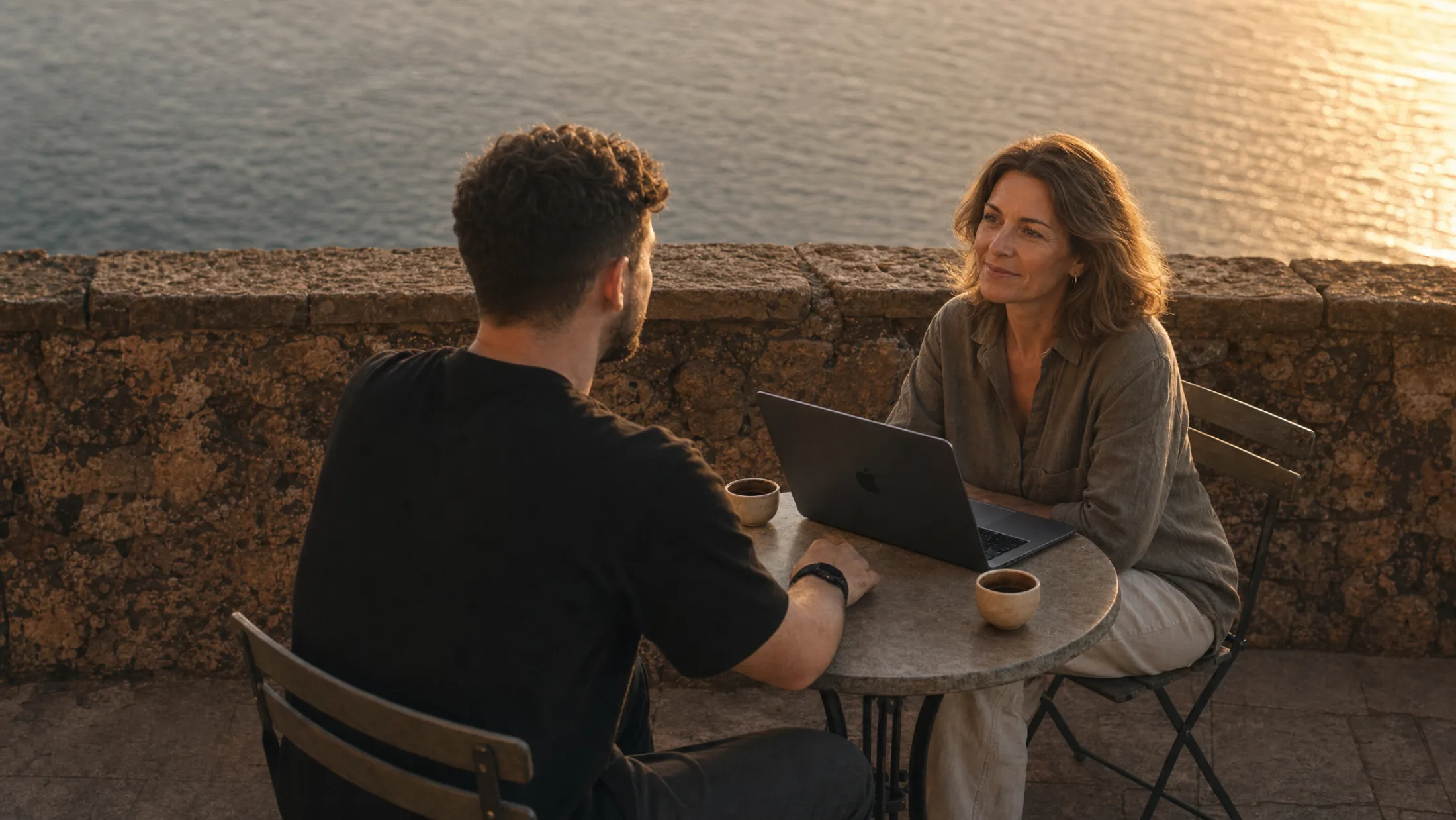 Daniel, seen from behind, in conversation with a woman at a small table with a laptop on a stone terrace above the sea