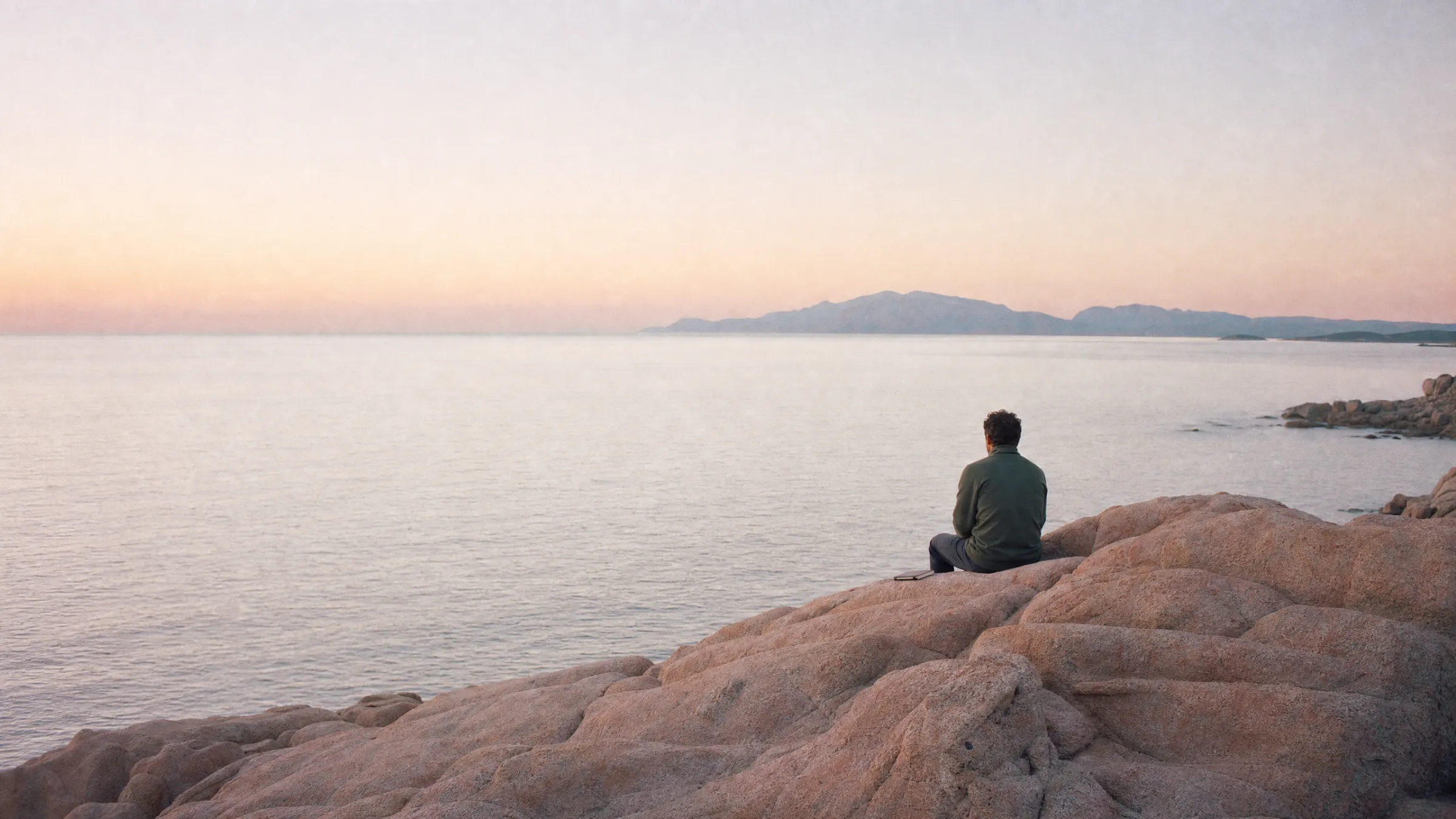 A calm sea at first light seen from pink granite rocks, a person sitting small on the rocks